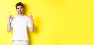 Komik olmayan yakışıklı adam kaşlarını çatıyor, kötü bir şeyi işaret ediyor, sarı arka plana şüpheyle bakıyor..