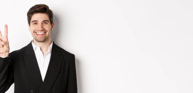 Siyah takım elbiseli yakışıklı iş adamının yakın çekimi, şaşkınlıkla gülümsüyor, iki numarayı gösteriyor, beyaz arka planda duruyor..