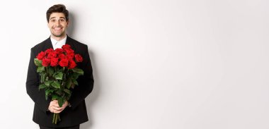 Siyah takım elbiseli yakışıklı bir adam resmi, elinde bir buket gül, gülümsüyor, beyaz arka planda duruyor..
