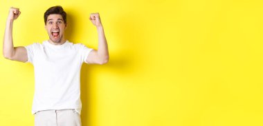 Heyecanlı bir adamın kazandığı, elini kaldırdığı ve kutladığı zafer ve takım için tezahürat yaptığı, sarı arka planda dikildiği bir görüntü. Boşluğu kopyala