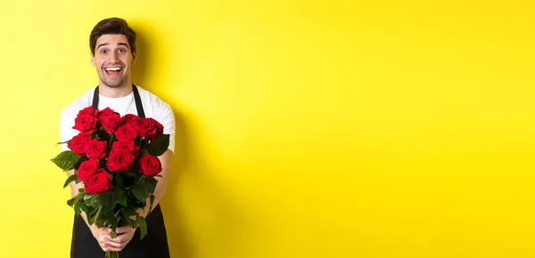 Seller in flower shop wearing black apron, giving bouquet of roses and smiling, standing over yellow background.