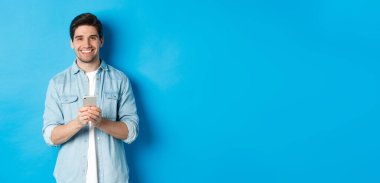 Sıradan kıyafetli yakışıklı adam kameraya gülümsüyor, akıllı telefonu kontrol ediyor, mavi arka planda duruyor..