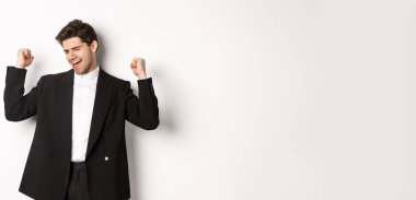 Başarılı ve memnun takım elbiseli yakışıklı bir adamın görüntüsü, sevinen ve yumruk sallayan işaretler yapan, mutluluktan dans eden, beyaz arka planda duran.