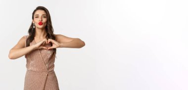 Kutlama ve parti konsepti. Şık giyinmiş güzel bir kadın, kırmızı dudaklar, kalp işareti gösteriyor ve öpücüğü bekliyor, beyaz arka planda duruyor..