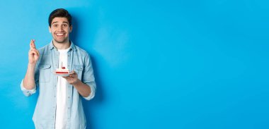 Umut dolu bir adam doğum günü pastası ve şans diliyor, dilek tutuyor, mavi arka planda duruyor..