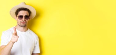 Turizm ve tatil kavramı. Yazlık şapkalı, güneş gözlüklü, sarı arka planda dikilip kamerayı işaret eden havalı adamın yakın çekimi..