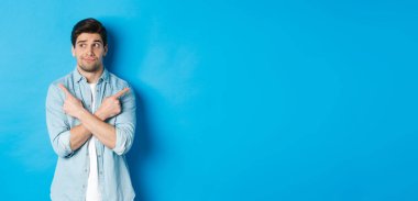 Kararsız yetişkin bir adamın portresi parmaklarını yana doğru gösteriyor ama sola bakıyor, iki ürün arasında seçim yapıyor, mavi arka plana karşı duruyor..