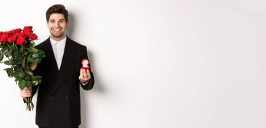 Siyah takım elbiseli yakışıklı, elinde güller ve nişan yüzüğü olan, onunla evlenme teklifi eden, beyaz arka planda duran....
