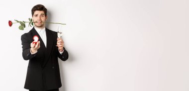 Takım elbiseli romantik genç bir adam evlenme teklif ediyor, elinde gül, bir kadeh şampanya, nişan yüzüğünü gösteriyor, evlenme teklif ediyor, beyaz arka planda duruyor..