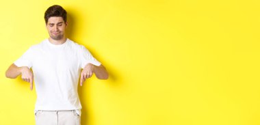 Beyaz tişörtlü şüpheci genç adam, üzgün ve aşağı bakıyor, onaylamıyor ve beğenmiyor, sarı arka planda duruyor.