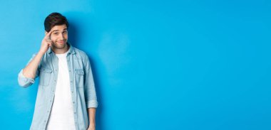 Genç düşünceli bir adam kafasını işaret ediyor, düşünmek istiyor, ipucu veriyor, mavi arka planda günlük kıyafetlerle dikiliyor..