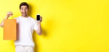 Alış veriş çantası tutan ve akıllı telefon ekranı, mobil bankacılık ve uygulama başarıları konsepti gösteren şaşırmış bir adam, sarı arka plan.