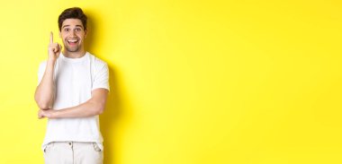 Çekici bir adamın fikri olduğunu hayal etmek, parmağını kaldırıp plan önermek, heyecanlı gülümsemek, sarı arka planda dikilmek..