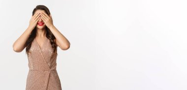 Kutlama, tatil ve parti kavramı. Cazip elbiseli çekici bir kadın Noel hediyesini bekliyor, gözleri ellerle örtüyor ve gülümsüyor, şaşırmayı bekliyor, beyaz arka plan..