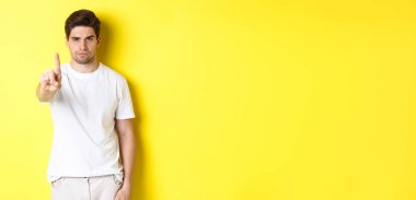 Hayır diyen kendine güvenen bir adam, bir şeyi reddeder ve yasaklar, bir parmağını gösterir ve kaşlarını çatar, sarı arka planda durur..