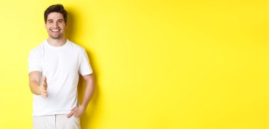 Yakışıklı ve kendine güvenen bir adam el sıkışmak için elini uzatıyor, seni selamlıyor, merhaba diyor, sarı arka planda beyaz tişörtle dikiliyor..