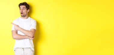 Beyaz tişörtlü etkileyici bir adam, sola bakıyor ve işaret ediyor, reklama bakın, sarı arka planda duruyor..