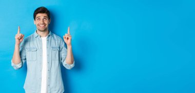 Sıradan giyinmiş yakışıklı bir adamın portresi. Mavi arka planda pankartı gösteriyor ve parmaklarını havaya kaldırıyor..