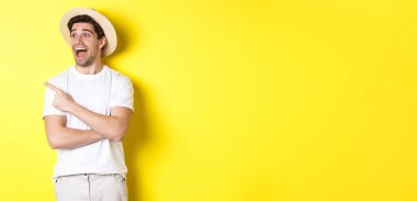 Turizm ve yaşam tarzı kavramı. Hasır şapkalı heyecanlı yakışıklı adam reklama bakıyor, sol üst köşedeki logoya bakıyor, sarı arka plan..