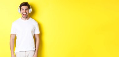 Kulaklıklı adam şaşırmış görünüyor. Beyaz elbiseli, sarı arka planda duruyor..