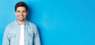 Kameraya gülümseyen, mavi arka planda sıradan kıyafetler giyen başarılı genç adamın yakın çekimi..