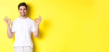 Rahatlamış adam gülümsüyor, tamam işaretleri gösteriyor, onaylıyor ya da kabul ediyor, sarı arka planda duruyor..