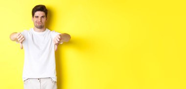 Şüpheci adam baş parmağını aşağı gösteriyor ve sırıtıyor, hoşlanmıyor ve katılmıyor, sarı arka planda duruyor. Boşluğu kopyala