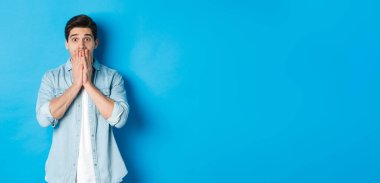 Şok olmuş yetişkin bir adamın görüntüsü kameraya hayranlıkla bakıyor, nefes nefese ve ağızlarını ellerle kapatıyor, mavi arka planda dikiliyor..