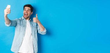 Mutlu adam selfie çekiyor ve mavi arka planda baş parmağını gösteriyor, elinde cep telefonu var..