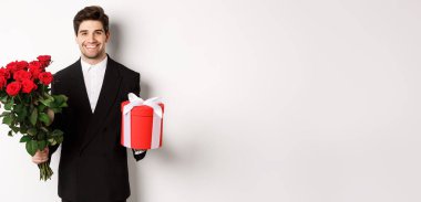 Tatil, ilişki ve kutlama kavramı. Siyah takım elbiseli yakışıklı erkek arkadaş, elinde bir buket kırmızı gül ve hediye, mutlu Noeller diliyor, beyaz arka planda duruyor..