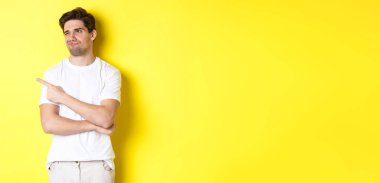 Etkilenmemiş genç adam şikayet ediyor, şüpheli bir ifadeyle kötü ürüne bakıyor, sarı arka planda duruyor..