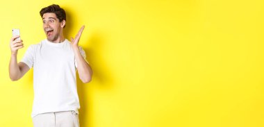 Şaşırmış ve mutlu bir adam cep telefonu ekranına bakıyor, harika haberler okuyor, sarı arka planda duruyor..