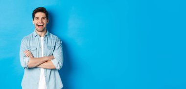 Sakallı, sıradan giyinmiş, komik bir şeye gülüp bakan, mavi arka planda duran yakışıklı bir adam..