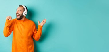 Mutlu adam kulaklıkla müzik dinlerken, elinde görünmez mikrofon tutarken, turkuaz arka planda dikilirken,.