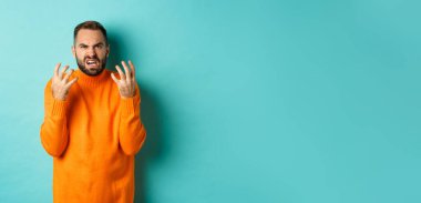 Hüsrana uğramış ve kızgın görünen öfkeli bir adamın görüntüsü, el sıkışıyor ve küçümseyerek yüzünü buruşturuyor, açık mavi arka planda sıkıntılı bir şekilde duruyor..