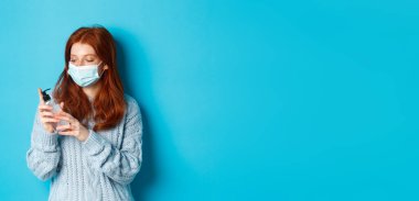 Kış, covid-19 ve sosyal uzaklık kavramı. Yüz maskeli genç kızıl kadın ellerini antiseptikle temizliyor, el dezenfektanı ile dezenfekte ediyor, mavi arka planda duruyor..