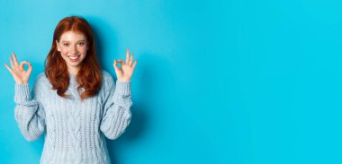 Kendine güvenen kızıl saçlı kız iyi işaretler gösteriyor, tercihi övüyor, onaylıyor ve onaylıyor, bir şey gibi, mavi arka planda duruyor. Boşluğu kopyala