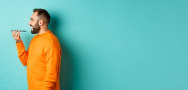 Yakışıklı sakallı bir adamın sesli mesaj kaydetme profili, cep telefonu tutmak ve hoparlörden konuşmak, mutlu bir şekilde gülümsemek, turkuaz arka planda durmak..