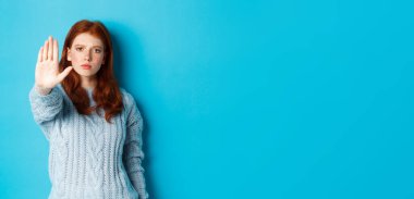 Ciddi ve kendine güvenen kızıl saçlı kız dur diyor, hayır diyor, hareketi yasaklamak için avucunu uzatıyor, mavi arka planda duruyor..