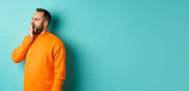 Yorgun sakallı bir adamın esneme fotoğrafı, ağzı elle kaplanmış, açık mavi arkaplana karşı uykulu duruyor..