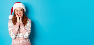 Kış ve Noel arifesi konsepti. Mutlu kızıl kız Noel 'i kutluyor, kameraya şaşkın şaşkın bakıyor, gülümsüyor ve yanaklarına dokunuyor, mavi arka planda duruyor..