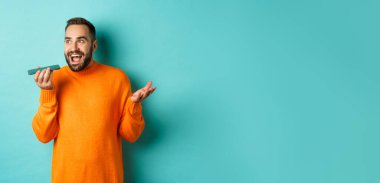 Hoparlörden konuşan mutlu bir adam, cep telefonuna işaret edip sesli mesaj kaydediyor, açık mavi arka planda turuncu süveter giyiyor..