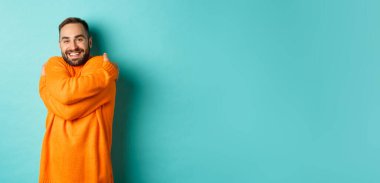 Mutlu adam kendini rahat hissediyor, sıcak bir süveter giyiyor ve kendine sarılıyor, gülümsüyor, açık mavi arka planda duruyor..