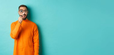 Neşeli adam bir şey arıyor, büyüteçten bakıyor ve gülümsüyor, mavi arka planda duruyor..