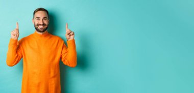 Memnun ve mutlu alıcı parmaklarını havaya kaldırarak gülümsüyor turkuaz arka planda turuncu süveter giyiyor..