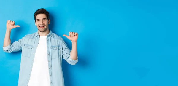 Portrait of confident man pointing at himself, being a professional, bragging while standing against blue background.
