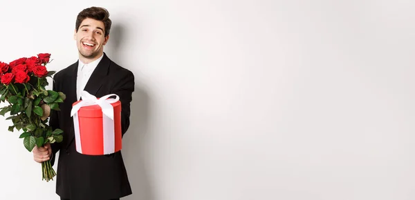 stock image Concept of holidays, relationship and celebration. Image of handsome smiling guy in black suit, holding bouquet of red roses and giving you a gift, standing against white background.
