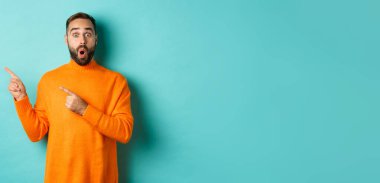 Sakallı, reklam pankartına tepki veren, solu gösteren, logoyu gösteren, şaşırmış görünen, turkuaz arka planda duran etkileyici genç adam..