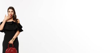 Güzellik ve moda konsepti. Siyah elbiseli, topuklu ayakkabı çantalı, kameraya bakan, şaşırmış, ağzı açık, beyaz arka planı olan bir kadın..