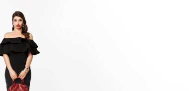 Güzellik ve moda konsepti. Uzun boyluysa, aptal genç kadın somurtuyorsa ve bakıyorsa kafası karışıksa, çanta tutuyorsa, topuklu giyiyorsa ve siyah elbise giyiyorsa, beyaz arka plan.